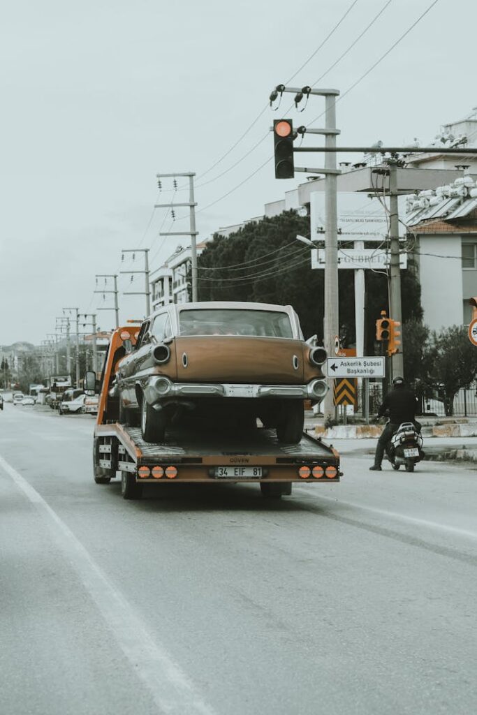 A vintage car is being towed through city streets on a tow truck, showcasing an urban environment.